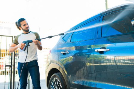 Téléchargez les photos : Jeune homme à sa maison garage laver et nettoyer sa voiture avec un pistolet à eau à l'extérieur - en image libre de droit