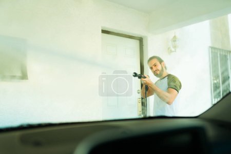 Téléchargez les photos : Latin jeune homme nettoyer son pare-brise et laver sa voiture avec un pistolet à eau - en image libre de droit