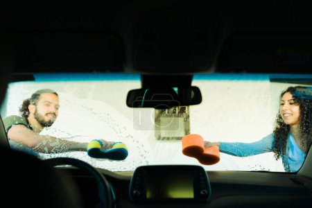 Téléchargez les photos : Couple heureux nettoyer le pare-brise avec du savon et une éponge tout en lavant la voiture ensemble - en image libre de droit
