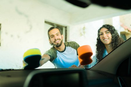 Téléchargez les photos : Couple heureux souriant regardant la caméra tout en nettoyant le pare-brise de la voiture à la maison - en image libre de droit