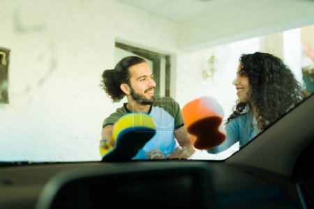 Foto de Hermosa pareja con el pelo rizado que se divierten lavando su coche con una esponja y limpiando el parabrisas - Imagen libre de derechos