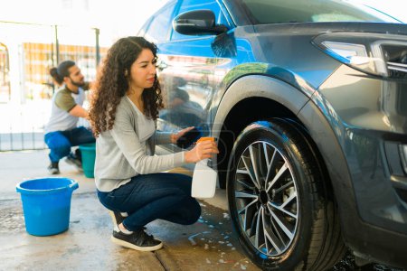 Foto de Mujer hispana brillando los neumáticos con un spray de producto mientras hace lavado de autos afuera - Imagen libre de derechos
