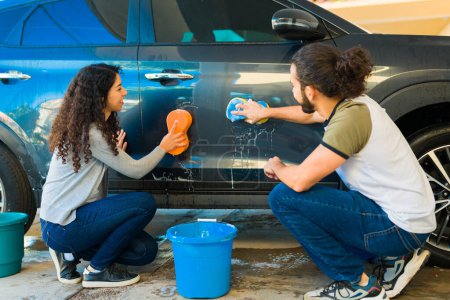 Téléchargez les photos : Vue arrière d'un couple heureux utilisant du savon et une éponge et faisant des tâches de lavage de voiture dans le garage - en image libre de droit