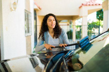 Téléchargez les photos : Belle femme heureuse souriant en utilisant un essuie-glace tout en nettoyant et en lavant sa voiture - en image libre de droit