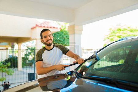 Foto de Hombre latino atractivo usando un limpiaparabrisas para limpiar el parabrisas después de hacer las tareas de lavado de autos - Imagen libre de derechos