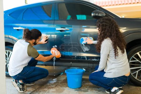 Foto de Pareja con el pelo rizado visto por detrás lavando su coche con jabón y una esponja con un cubo - Imagen libre de derechos