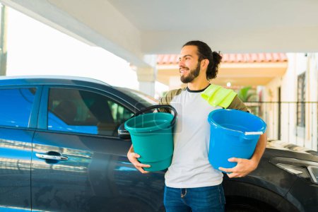 Téléchargez les photos : Heureux homme hispanique souriant tout en portant des seaux avec de l'eau et du savon finissant de laver la voiture - en image libre de droit