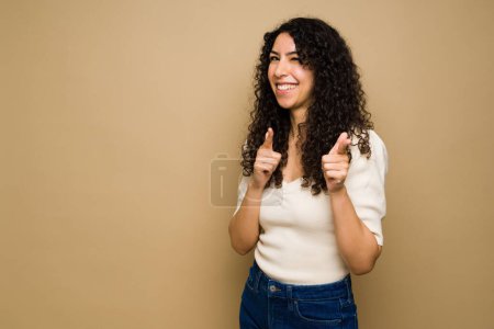 Foto de Cheerful young woman pointing to you and doing finger guns while smiling in front of a yellow studio background - Imagen libre de derechos