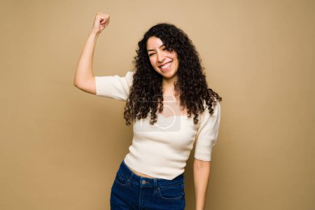 Foto de Attractive happy latin young woman smiling and showing her bicep while feeling strong and powerful - Imagen libre de derechos