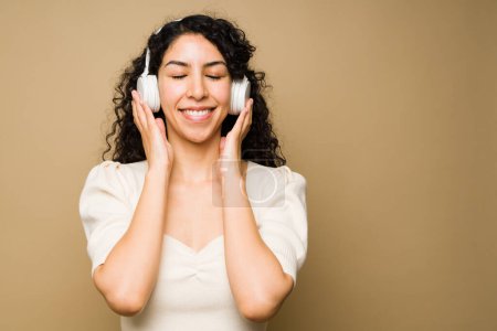 Téléchargez les photos : Cheerful young woman listening to music with headphones closing her eyes and relaxing against a studio background with copy space - en image libre de droit