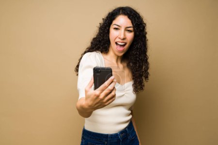 Photo for Surprised hispanic woman looking shocked while looking at a text or social media on her smartphone - Royalty Free Image