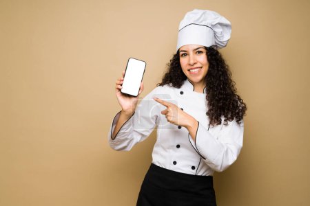 Photo for Attractive woman chef or cook smiling while pointing to her smartphone for a food delivery app or social media - Royalty Free Image
