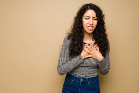 Stressed latin young woman having chest pain or suffering a heart attack against a studio background