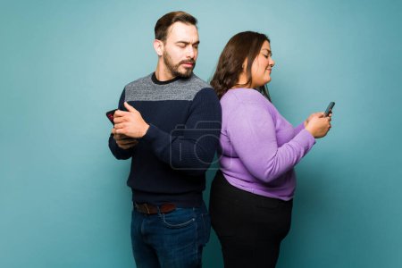 Photo for Upset caucasian man spying on his latin girlfriend texting another man on the smartphone while cheating - Royalty Free Image