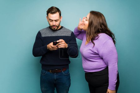 Foto de Angry overweight woman yelling to the ear of her annoyed caucasian boyfriend texting on the smartphone while fighting - Imagen libre de derechos