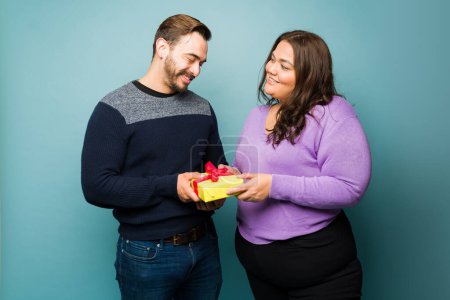 Photo for Beautiful big woman smiling giving an anniversary present to her happy caucasian boyfriend against a studio background - Royalty Free Image