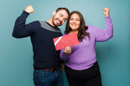 Foto de Excited young couple doing a bicep curl and feeling strong and powerful while looking happy holding a gift card - Imagen libre de derechos