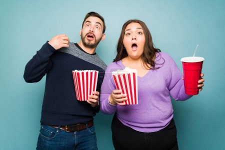 Foto de Surprised young couple looking shocked while watching a movie and eating popcorn - Imagen libre de derechos