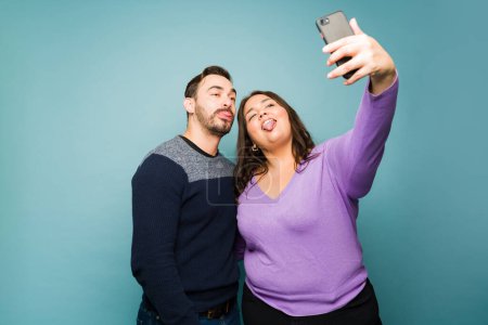 Photo for Caucasian boyfriend and overweight girlfriend doing funny faces while taking selfies for social media with a smartphone - Royalty Free Image