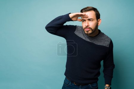 Photo pour Attractive young man putting a hand above his eyes and squinting while looking for something in the distance and looking far away - image libre de droit