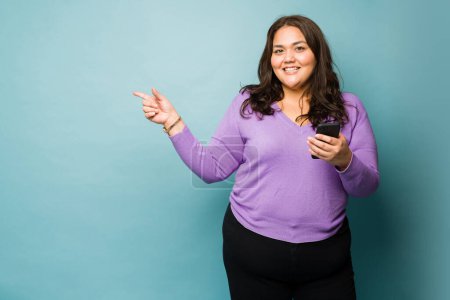 Téléchargez les photos : Attractive latin obese woman using her smartphone and pointing with a finger to copy space against a studio background - en image libre de droit