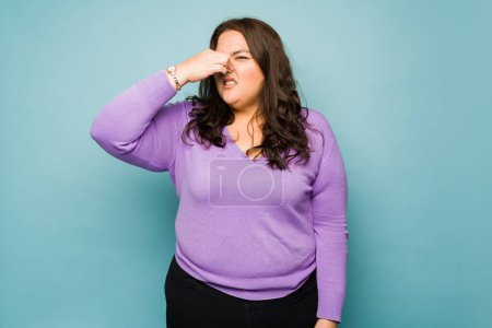 Photo for Annoyed upset overweight woman covering her nose because of bad smell against a studio background - Royalty Free Image