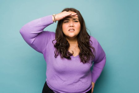 Téléchargez les photos : Overweight young woman squinting and putting a hand above her eyes trying to find or looking ahead - en image libre de droit