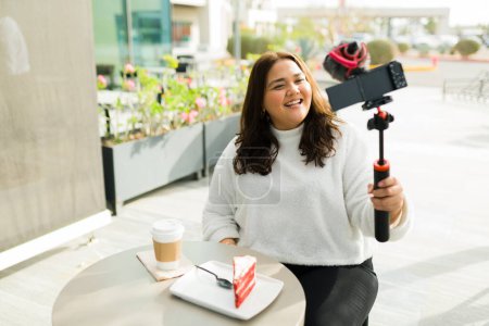 Photo for Cheerful beautiful hispanic woman working as an influencer filming a video with a camera at the coffee shop for her social media content - Royalty Free Image