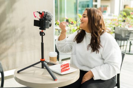 Photo for Chubby latin influencer woman doing a restaurant or cafe review for social media while filming video content with a camera - Royalty Free Image