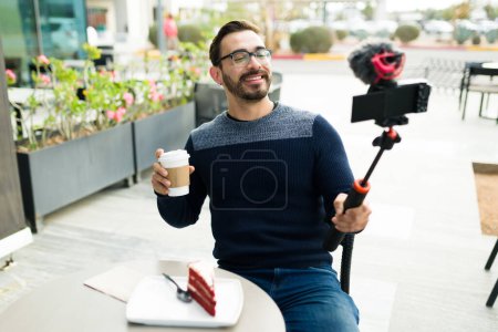 Photo for Cheerful caucasian man influencer smiling filming with a camera and drinking coffee while eating cake at a cafe - Royalty Free Image