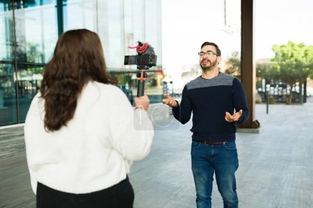 Photo for Large young woman filming a caucasian man influencer talking to the camera outdoors in the city for his social media - Royalty Free Image