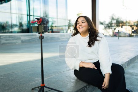 Photo for Cheerful large woman using a camera and a tripod to film a video blog to make social media content in the city - Royalty Free Image