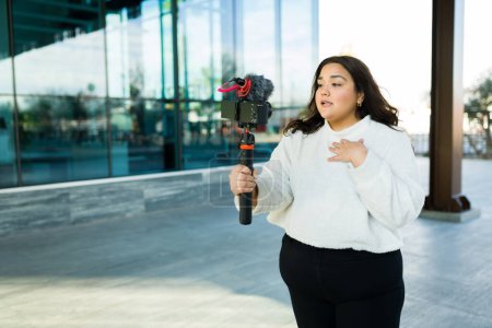 Photo for Attractive big woman using a camera and stabilizer to film a video blog and making social media content - Royalty Free Image