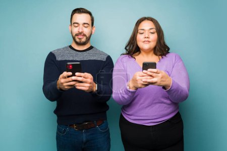 Photo for Distracted young couple texting and using social media on the smartphone against a blue studio background - Royalty Free Image