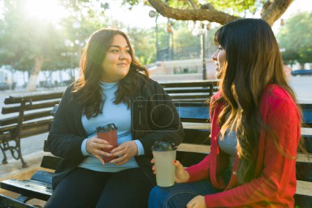 Téléchargez les photos : Hispanic young women friends talking while hanging out in the park and enjoying a cold drink together - en image libre de droit