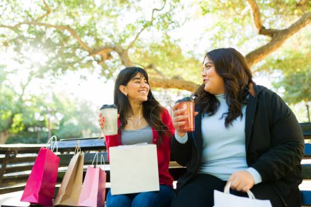 Foto de Attractive hispanic friends drinking coffee and smiling at the park after going shopping - Imagen libre de derechos