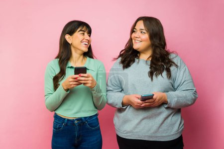 Foto de Smiling young women friends looking excited laughing while texting on their smartphones in front of a pink background - Imagen libre de derechos