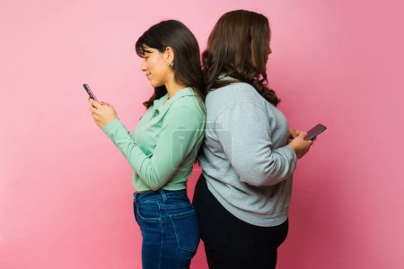 Foto de Profile of distracted hispanic young women standing back to back texting on their smartphones - Imagen libre de derechos