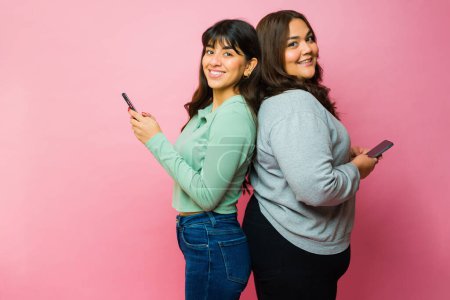 Foto de Portrait of happy women best friends smiling while making eye contact and texting on the smartphone - Imagen libre de derechos