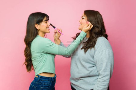 Foto de Beautiful latin women friends putting on makeup with a brush and eyeliner getting ready to go out together - Imagen libre de derechos