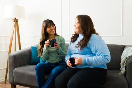 Photo for Happy beautiful young women and best friends talking drinking coffee while hanging out together at home - Royalty Free Image
