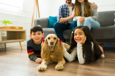 Foto de Smiling little boy and girl smiling and making eye contact while petting their beautiful yellow cocker spaniel dog in the living room - Imagen libre de derechos