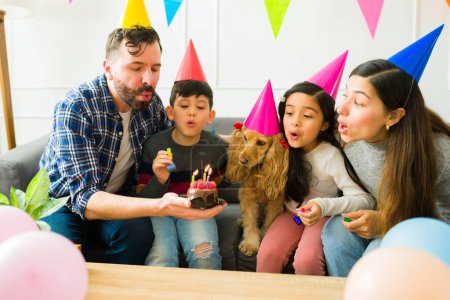 Foto de Happy parents with their young kids blowing the cake candles while celebrating a birthday party together at home - Imagen libre de derechos