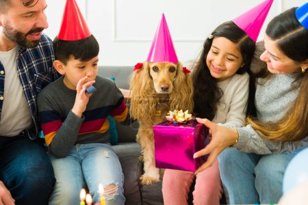 Foto de Cute yellow cocker spaniel dog with a party hat receiving birthday presents from its family and kids - Imagen libre de derechos