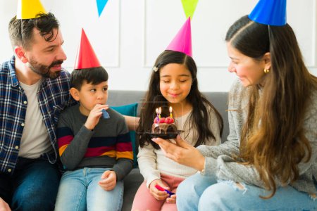 Foto de Adorable hispanic girl making a wish and ready to blow the candles while celebrating her birthday with her little family - Imagen libre de derechos
