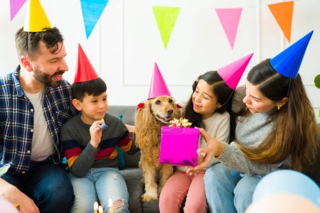Foto de Excited young kids with their parents giving presents to their cocker spaniel dog while celebrating a birthday party at home - Imagen libre de derechos