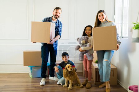 Foto de Portrait of a beautiful happy family with little kids and a dog smiling carrying boxes while moving to a new home - Imagen libre de derechos