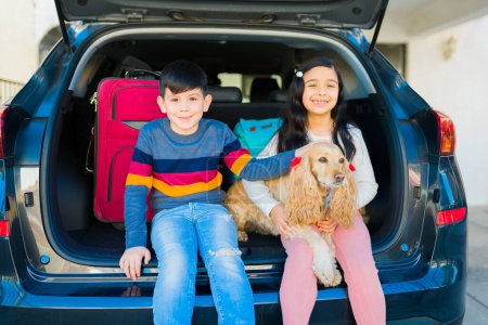 Téléchargez les photos : Adorable young children smiling while waiting with the suitcases in the car trunk to start their vacations with their cute dog - en image libre de droit