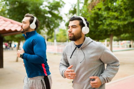 Téléchargez les photos : Hispanic young man and friend running and doing exercises wearing headphones outdoors - en image libre de droit