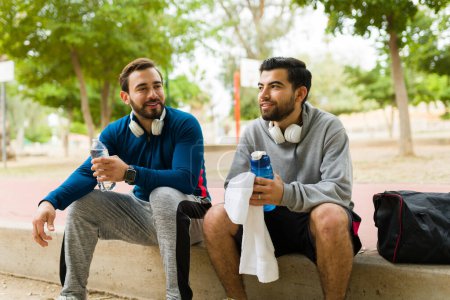 Téléchargez les photos : Attractive men friends smiling while talking drinking water after exercising and running at the park - en image libre de droit